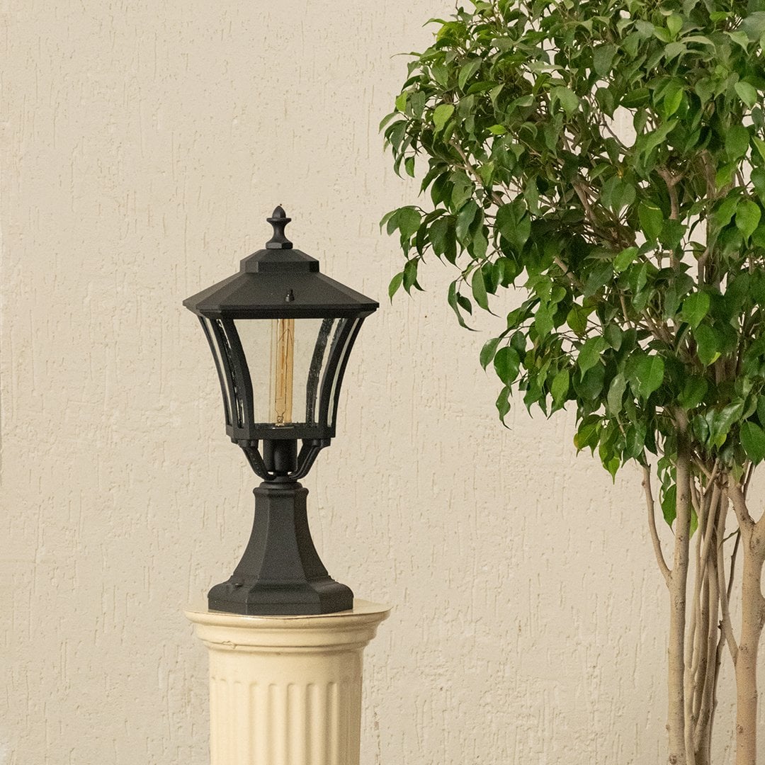 Black outdoor pillar lantern with light on placed on a decorative pedestal beside a green plant adding warm ambient glow and elegant exterior styling to the space.