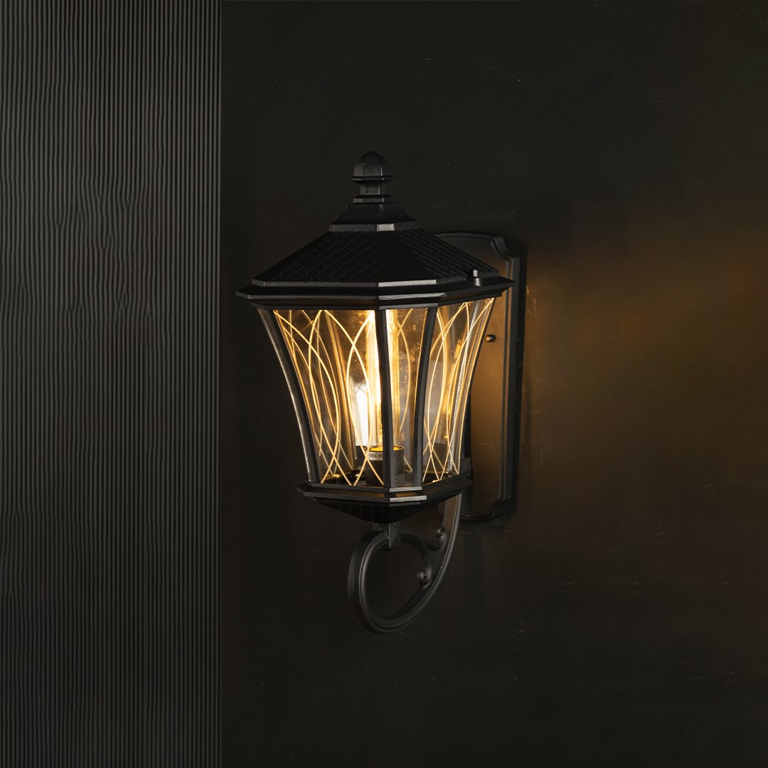 Black vintage wall lantern illuminated against a dark background showcasing its metal frame clear patterned glass and warm ambient light.