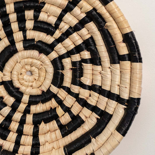 A closeup view of the natural and black rattan wall decor plate showing its geometric spiral weaving and textured handcrafted finish for warm earthy home styling.