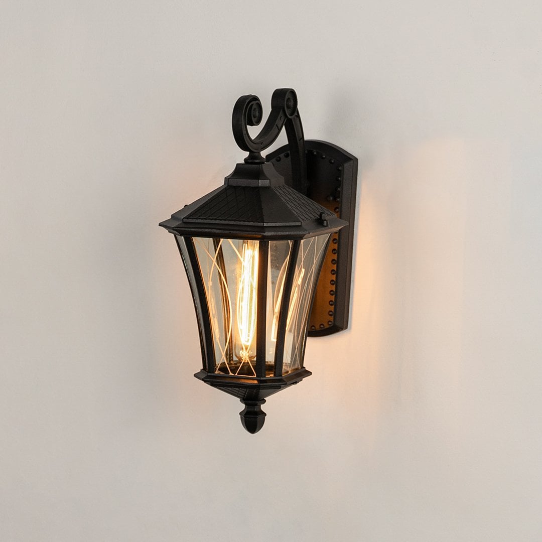 Black metal outdoor lantern with textured glass shown with light on against a white background highlighting its warm glow and traditional exterior design.