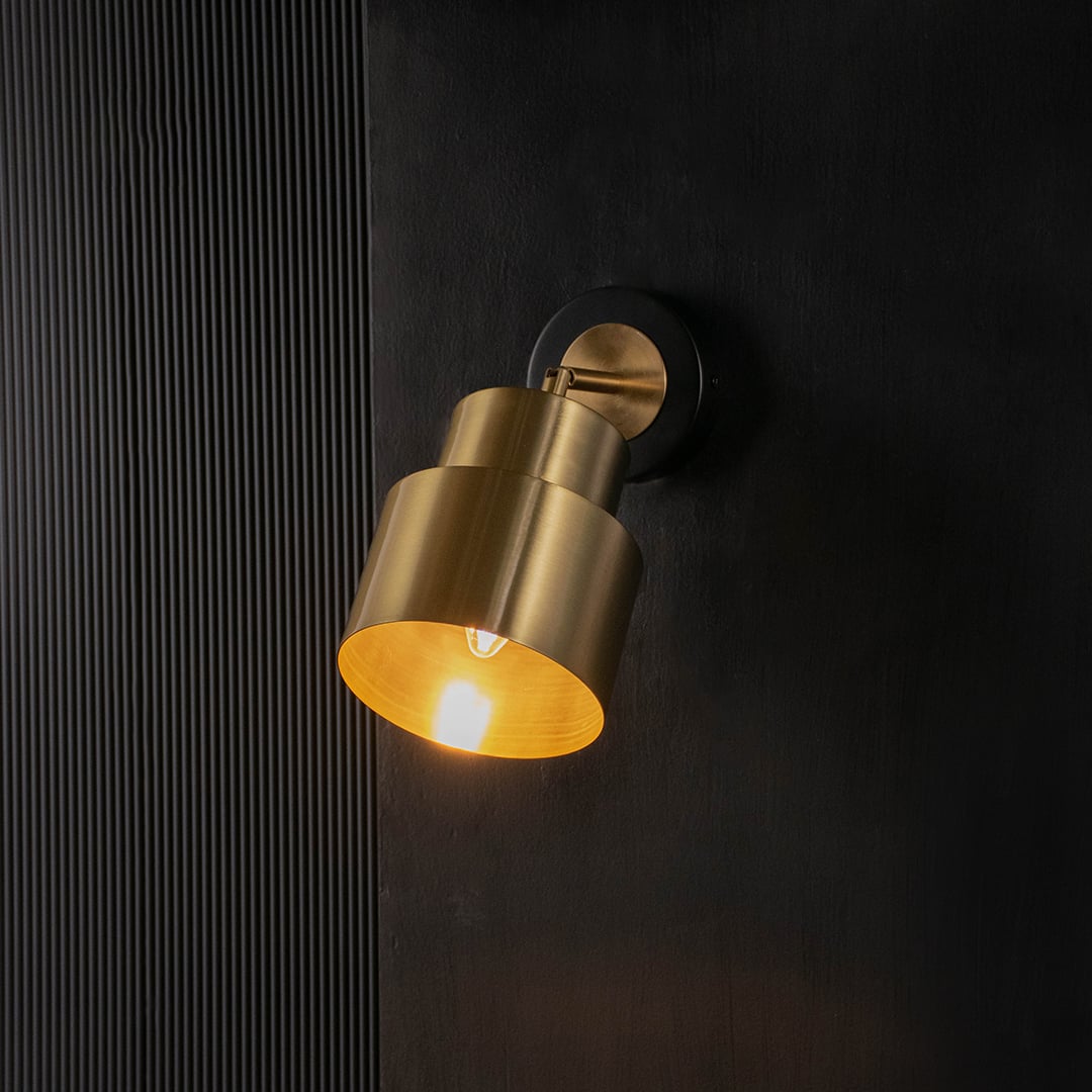 Gold wall light with a layered metal shade glowing softly against a black background highlights its modern industrial design.