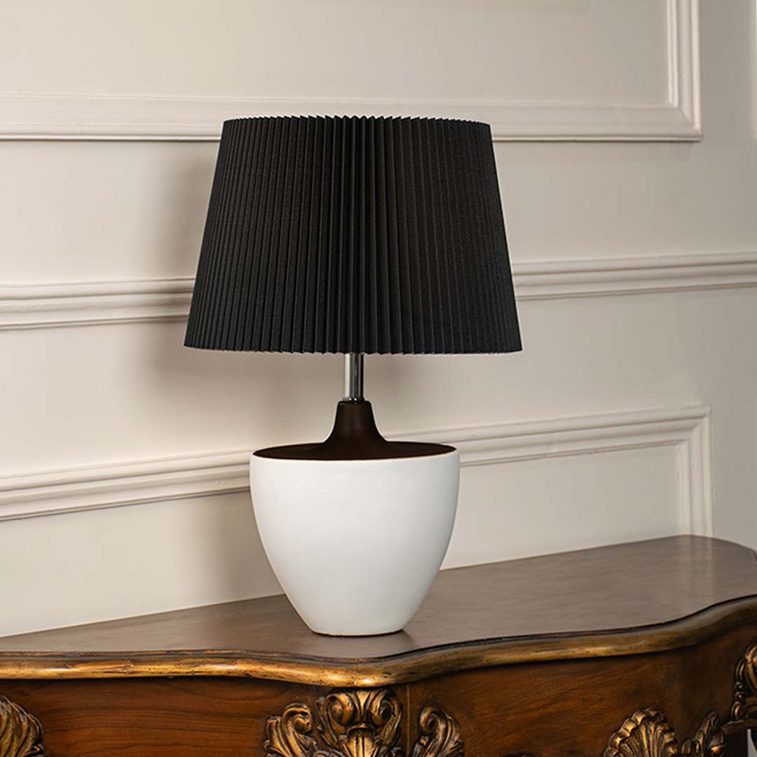 Black table lamp with pleated shade and white ceramic base lit on a clean white background for product display.