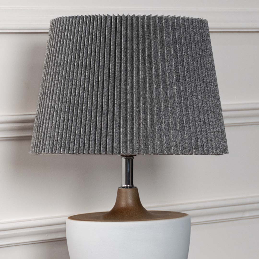 Closeup of white ceramic base table lamp with textured grey pleated shade detailing its modern simple form and refined craftsmanship.