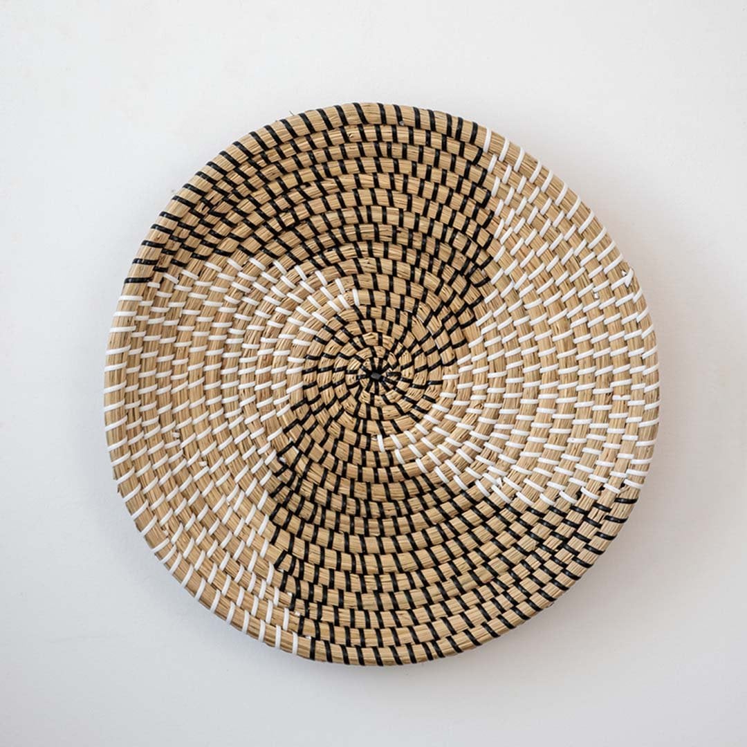 Handwoven wicker basket wall decor in beige with black and white spiral woven geometric pattern on clean white background.