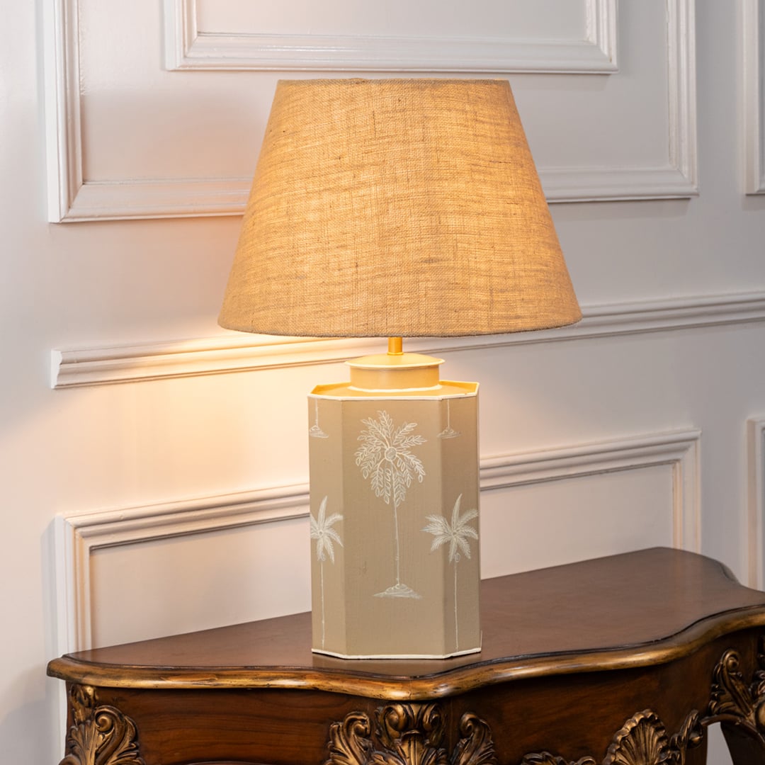 Palm design table lamp turned on with warm golden glow highlighting beige ceramic base with white palm motifs and jute textured shade ideal for living room lighting.