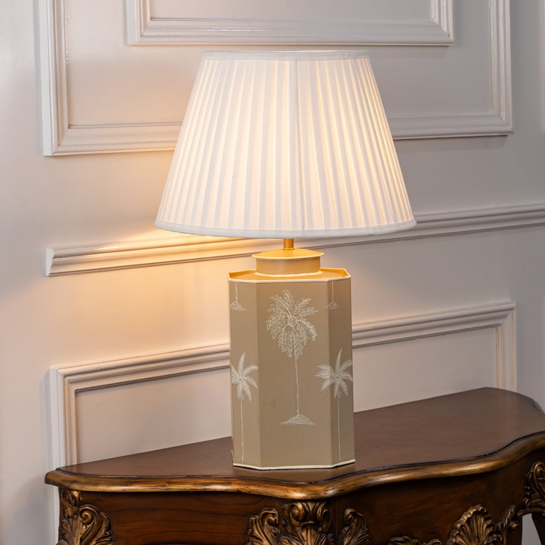 Beige palm table lamp turned on with warm white glow featuring hand-painted palm motifs and pleated white shade ideal for living room or bedside decor.