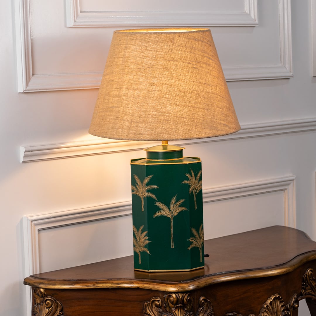 Green palm table lamp turned on with warm white light featuring hand-painted golden palm motifs and pleated white shade perfect for elegant living room lighting.