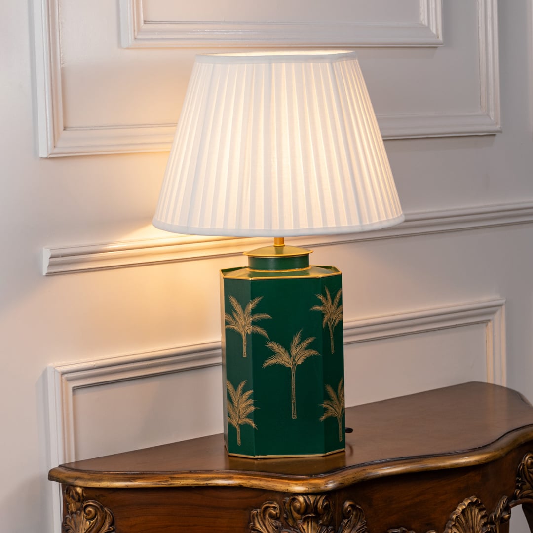 Green palm table lamp turned on with soft warm glow featuring dark green ceramic base with golden palm tree motifs and pleated white shade perfect for elegant home lighting.