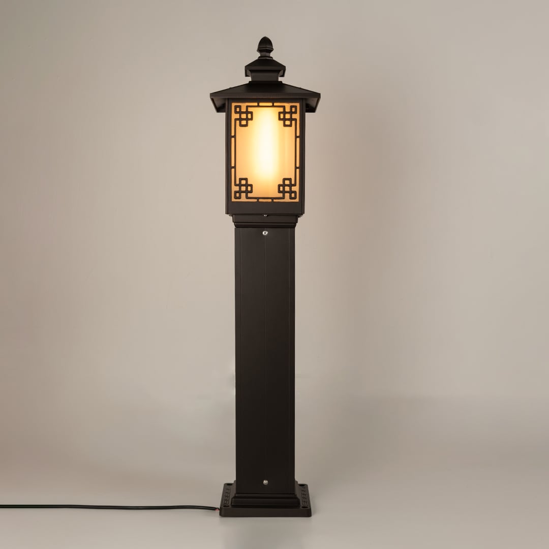 A black outdoor bollard lamp displayed on a white background with the light on highlighting its frosted glass panel and ornate geometric frame in a clean studio view.