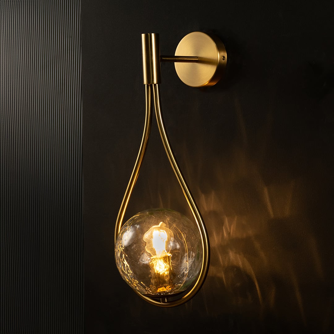 Brushed gold teardrop wall light glowing on black background creating warm dramatic illumination with elegant hammered glass globe design.