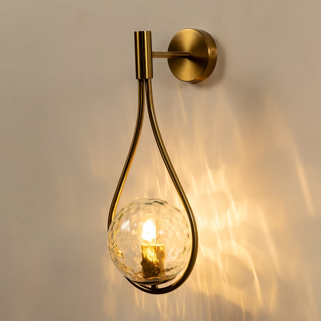 Brushed gold teardrop wall light glowing on white background showcasing warm illumination and artistic curved frame with hammered glass globe.