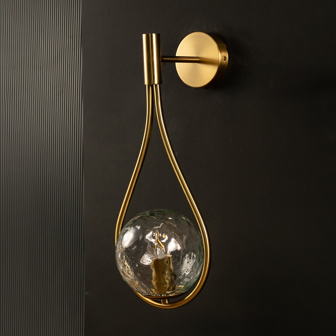 Brushed gold teardrop wall light with lights off on black background highlighting its artistic curved frame and clear hammered glass globe.