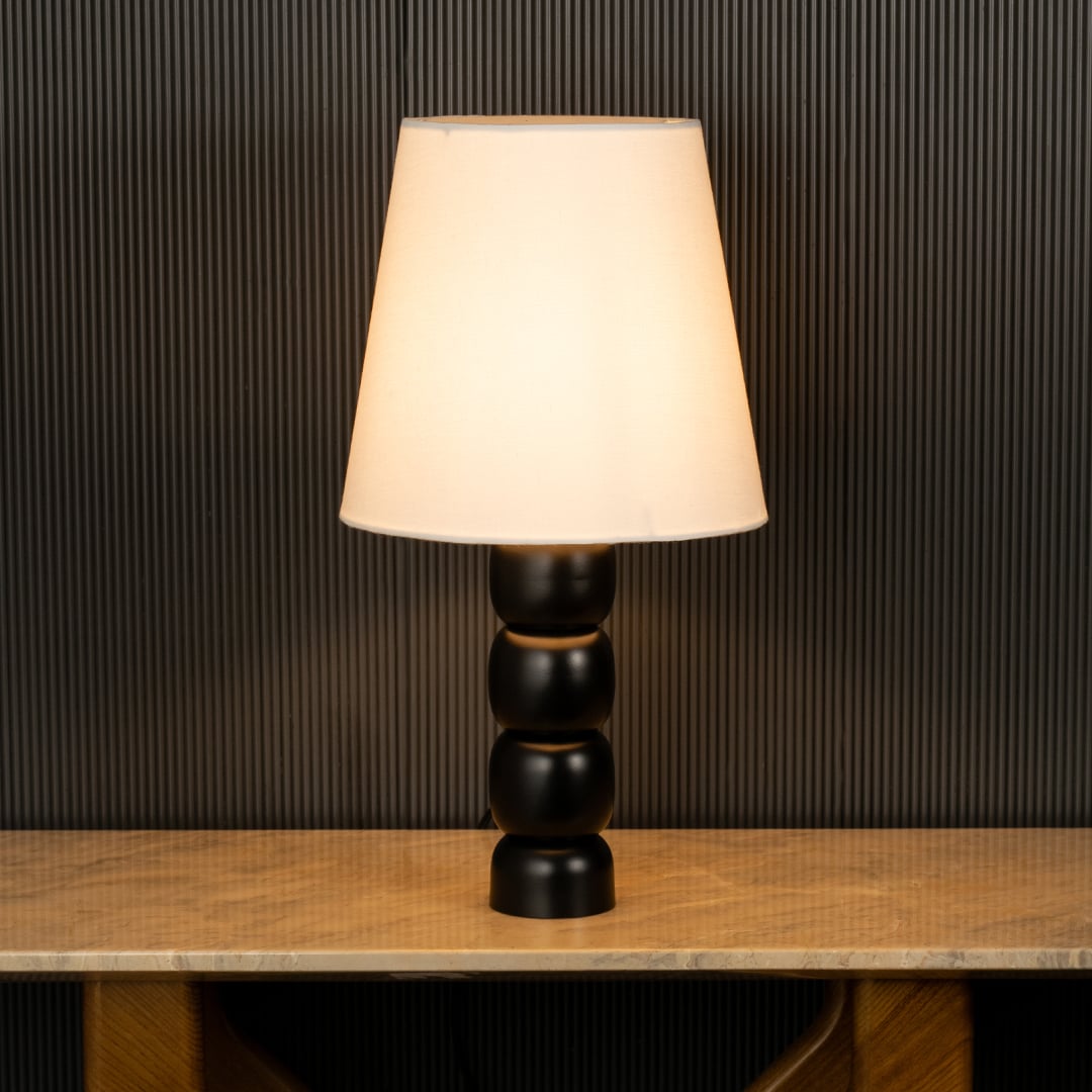 Table lamp with white shade and black stacked base casting warm ambient glow imagined against a dark background creating a dramatic modern interior decor mood.