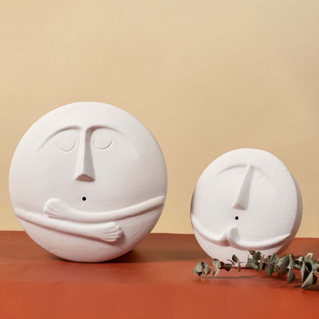 Set of two round white ceramic face vases in different sizes arranged together against a warm-toned background with decorative greenery.