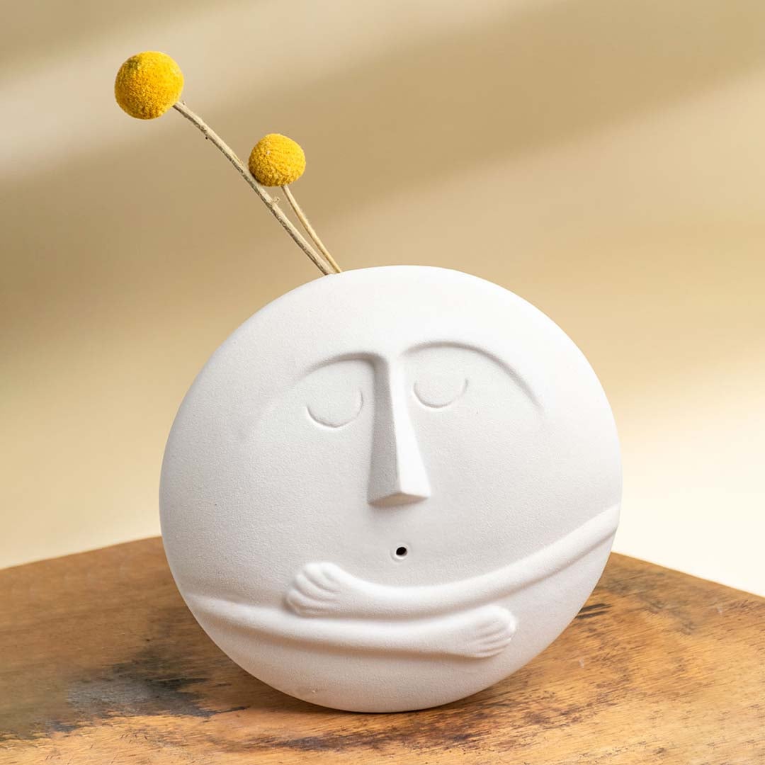 Decorative round white ceramic face vase displayed on a wooden surface with soft warm lighting and yellow billy ball stems adding a playful accent.