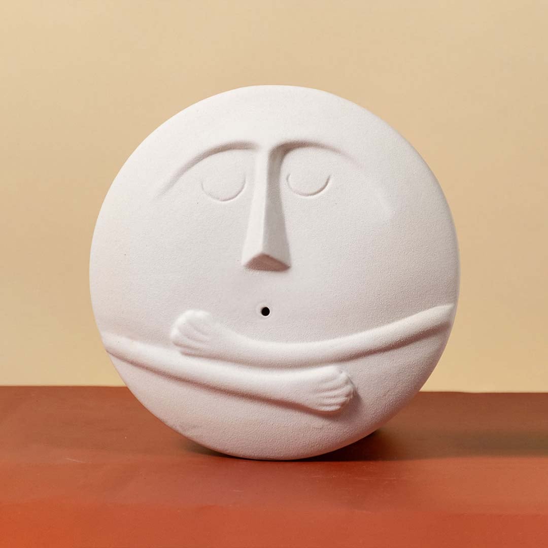 Round white ceramic face vase with embossed facial features and hugging arms showcased against a clean light background.