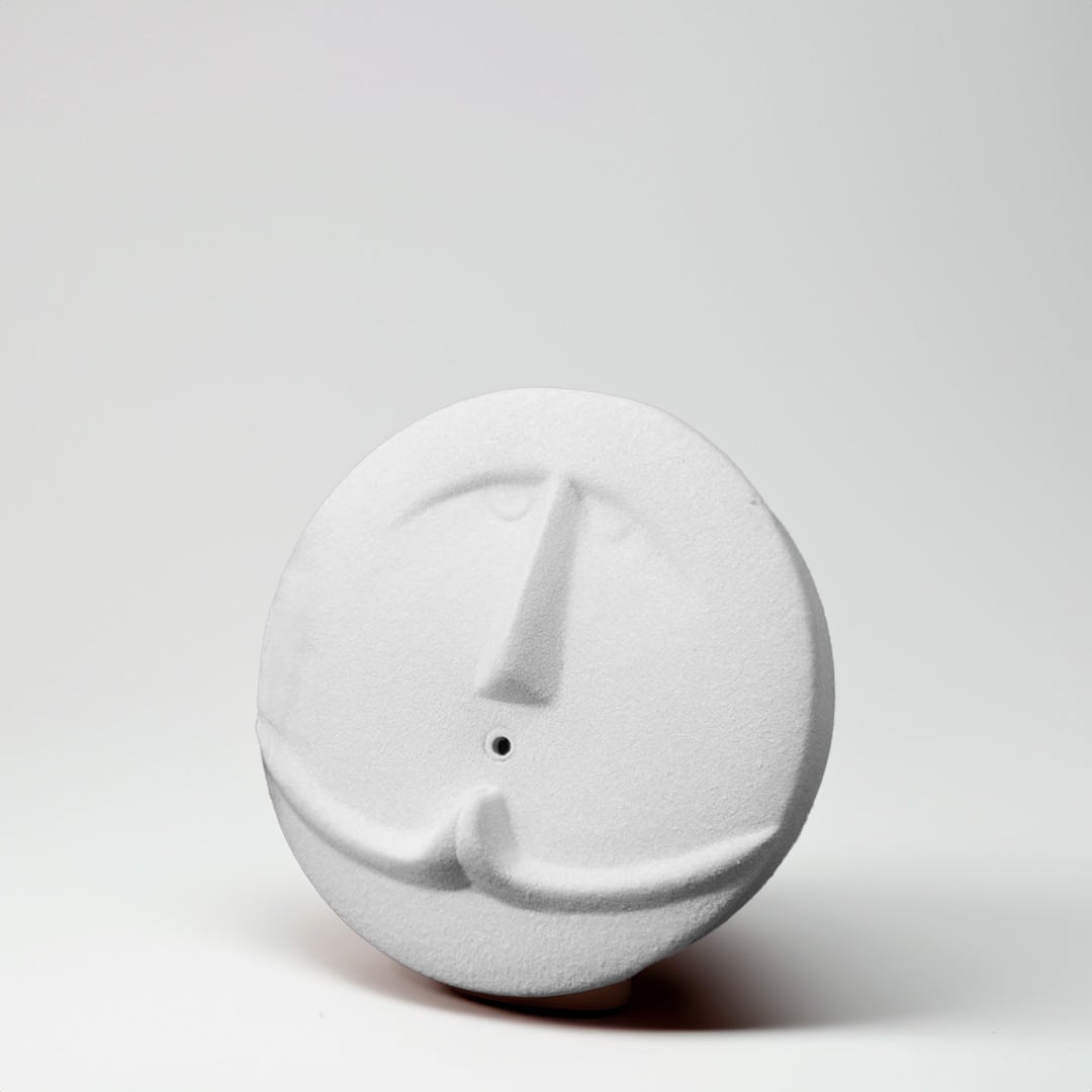 Small round white face sculpture vase placed on a clean pale surface with soft light enhancing its minimalist shape and carved features.