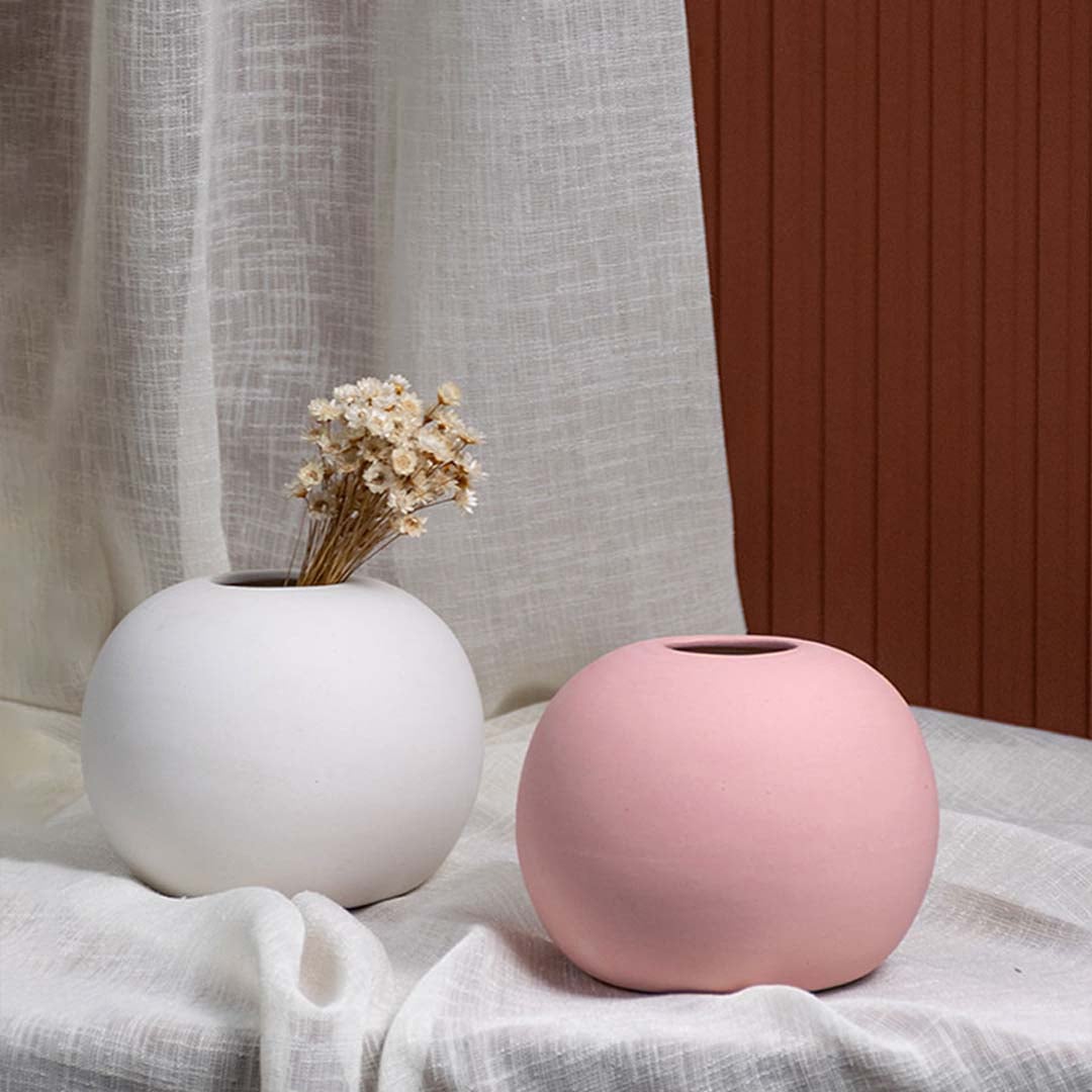 White ceramic round vase with dried flowers grouped with a pink vase on soft draped fabric creating a warm elegant decorative setup.