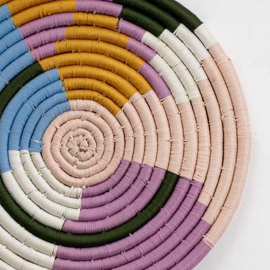 Closeup view of a handwoven round wall decor showing tightly coiled fibers in pastel blue mustard purple green and beige with detailed texture visible.