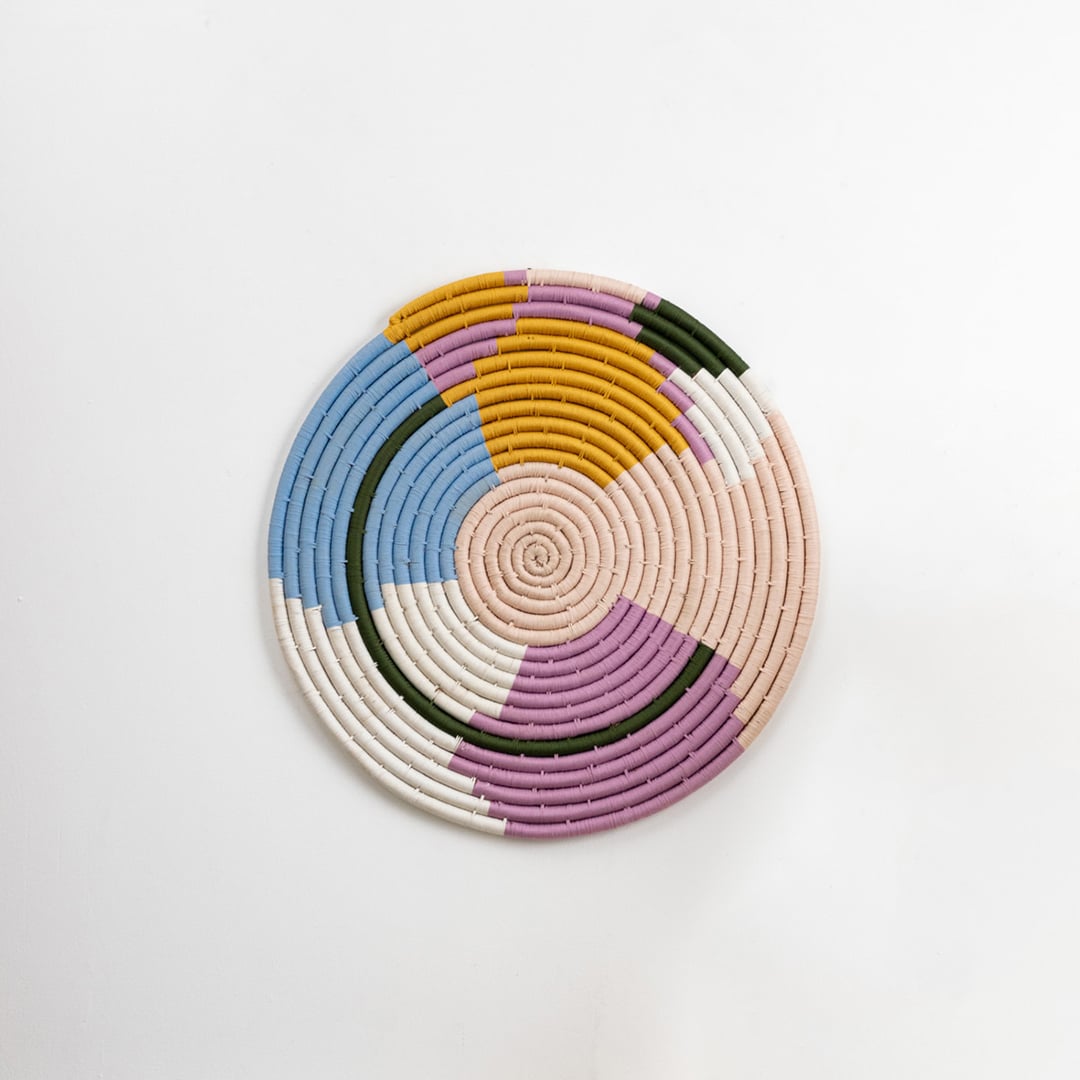 Round cotton woven wall plate in pastel blue pink mustard green and cream tones shown on a clean white background for modern artistic wall decor styling.
