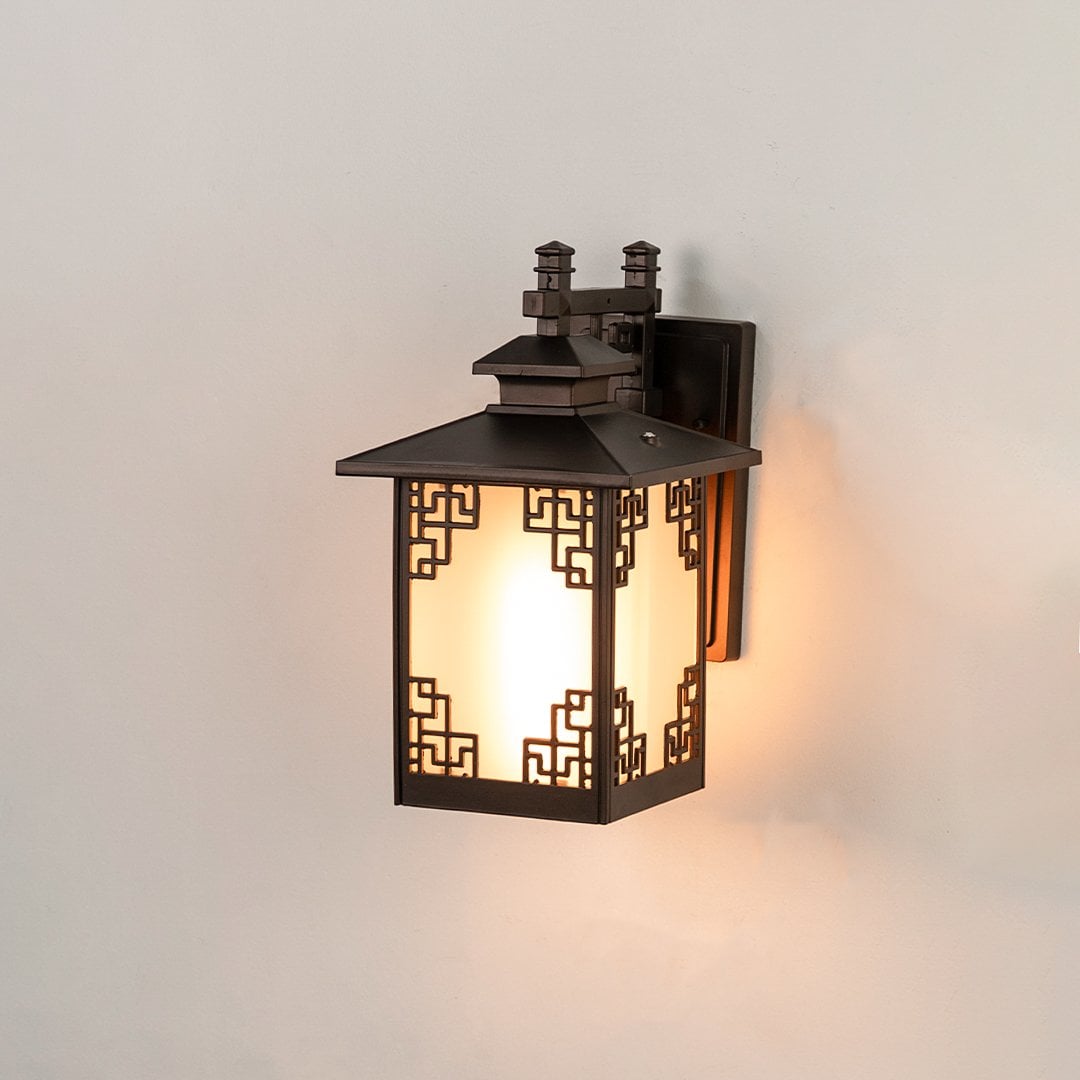 Brown metal outdoor lantern on white background with light on showing warm glow behind frosted glass and decorative geometric frame for elegant exterior lighting.
