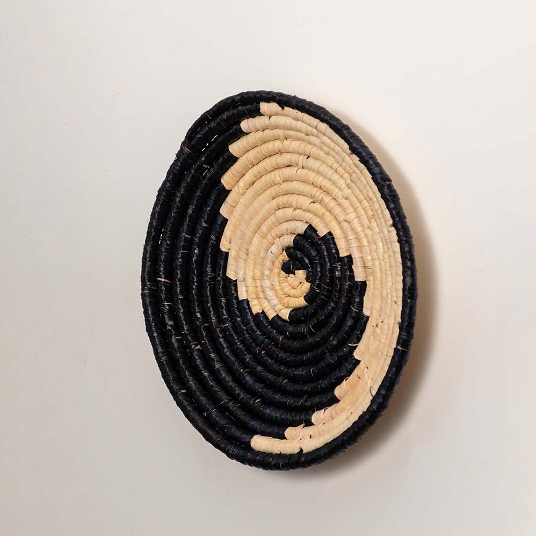 A side angle view of the handwoven natural and black rattan wall decor plate showing its curved rim and textured detailing for modern handcrafted home styling.