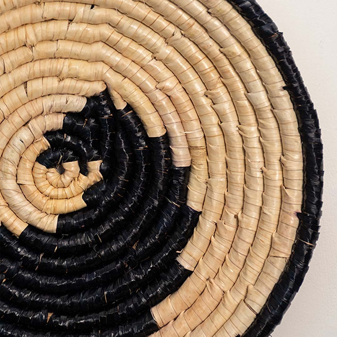 A closeup view of the natural and black rattan wall decor plate showing its spiral artisan weave and textured handcrafted detailing for warm earthy home styling.
