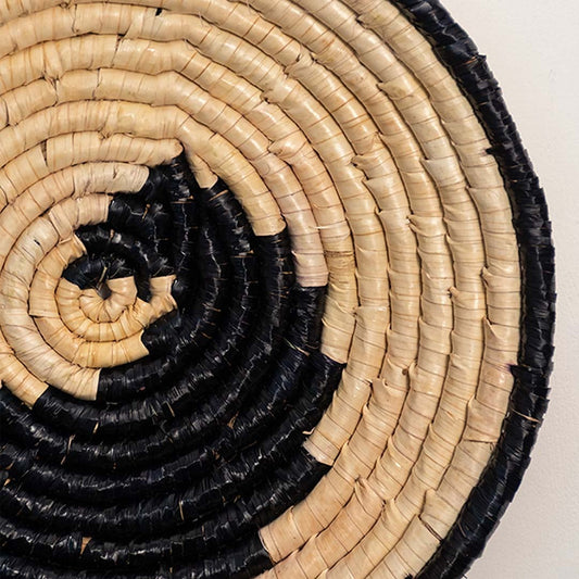 A closeup view of the natural and black rattan wall decor plate showing its spiral artisan weave and textured handcrafted detailing for warm earthy home styling.
