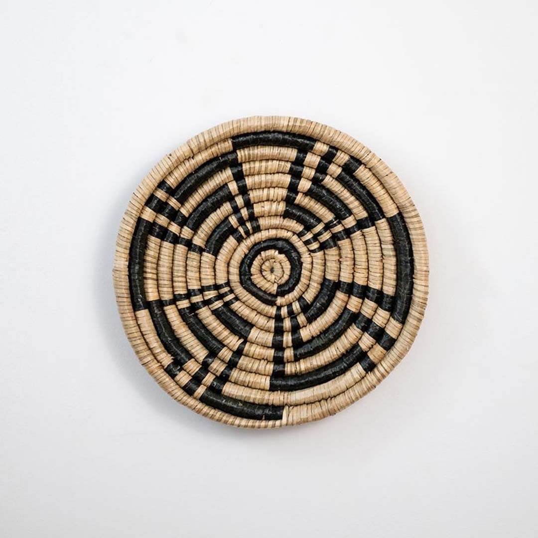 Round woven wall basket in natural beige and black seagrass pattern displayed on white background for wall decor styling.