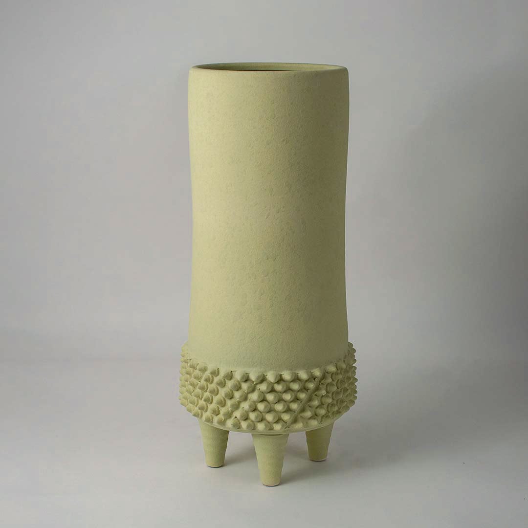 Matte light green ceramic vase with textured pattern and tripod legs on white background showing modern artistic decor.