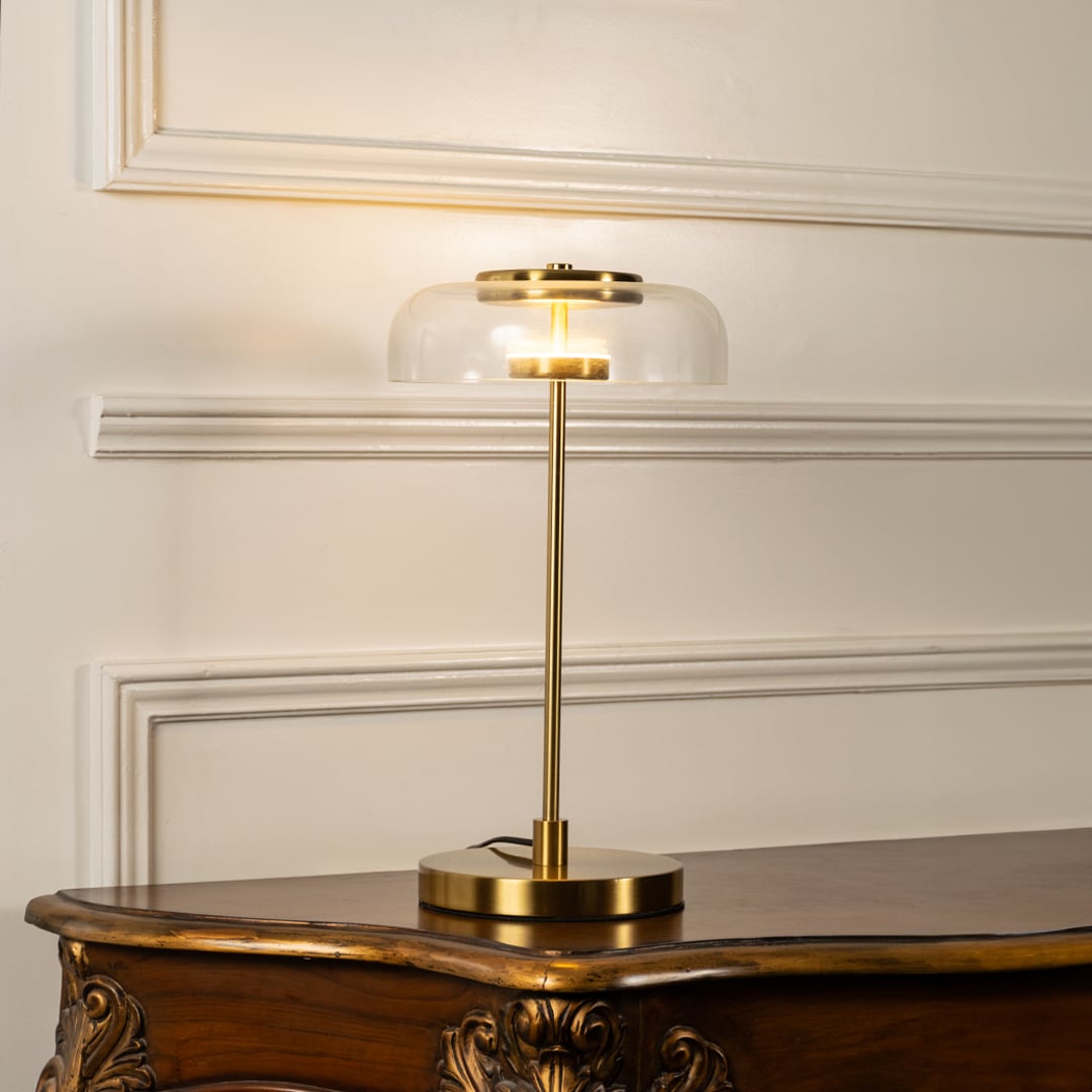 Gold table lamp with illuminated clear glass dome shade and slim metal stem shown on a white background ideal for bedside tables and modern decor.