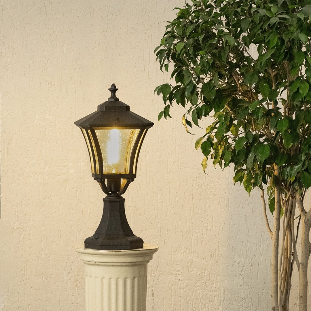 Black outdoor pillar lantern with light on placed on a decorative pedestal creating warm ambient glow with textured glass and classic metal frame for garden decor.