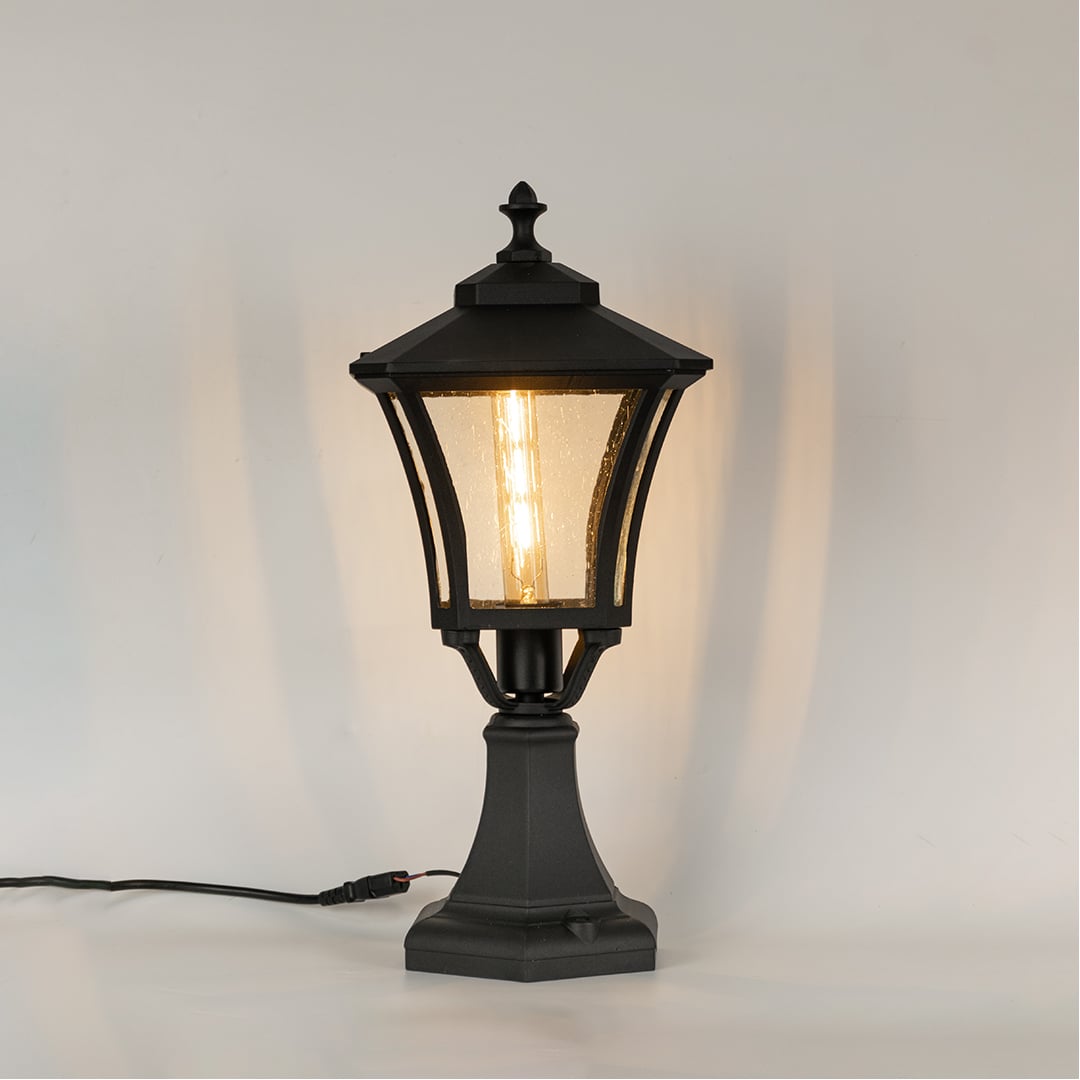 Black pillar lantern on white background with light on showing glowing textured glass and classic metal frame suitable for pathways patios and exterior decor.