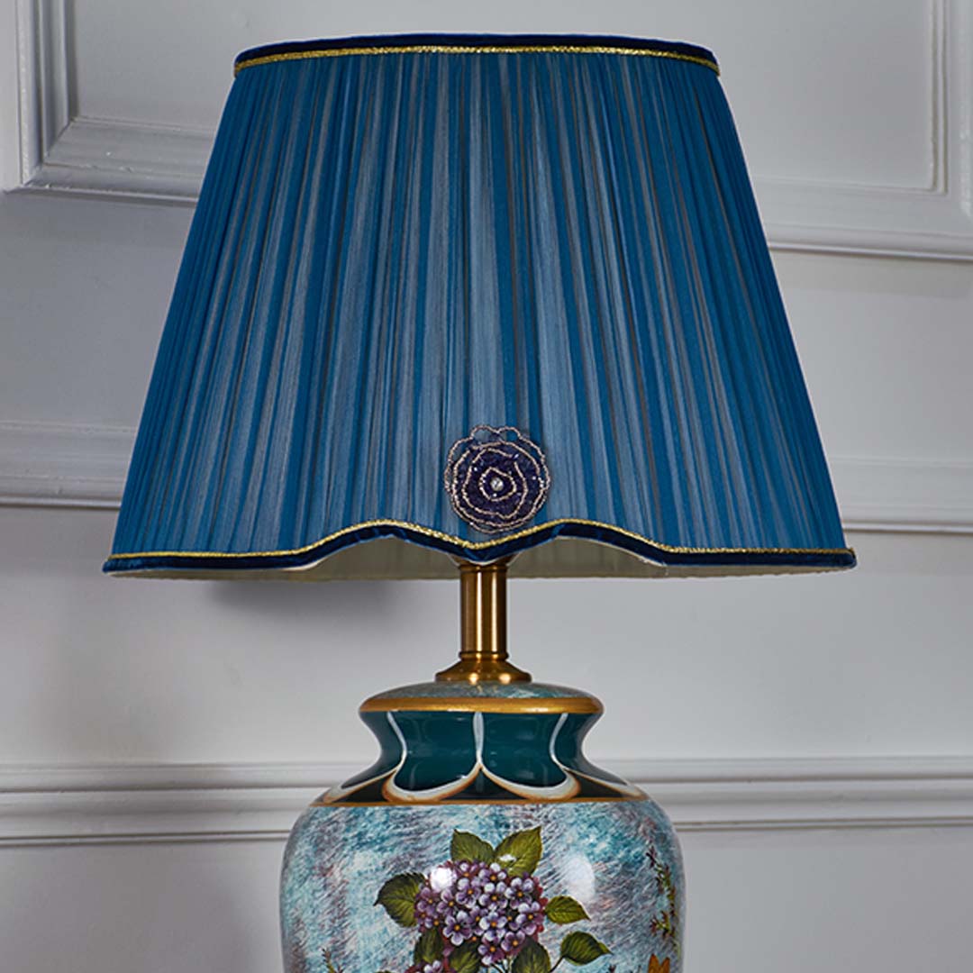 Closeup of blue ceramic table lamp with hand painted floral pattern gold base and textured pleated blue shade showcasing premium craftsmanship.