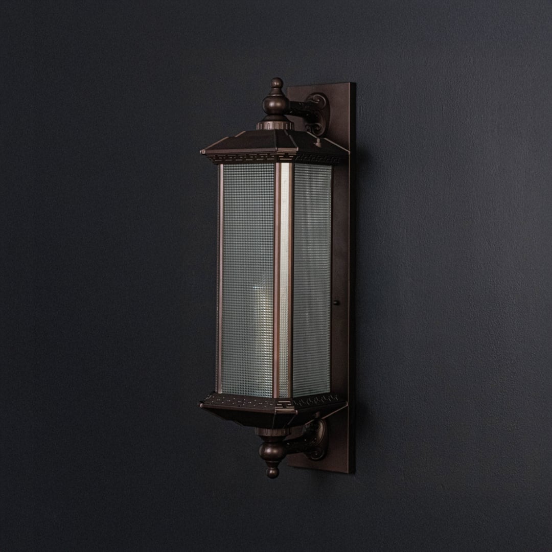 Bronze wall light with frosted glass body and ornate detailing displayed off against dark background surface.