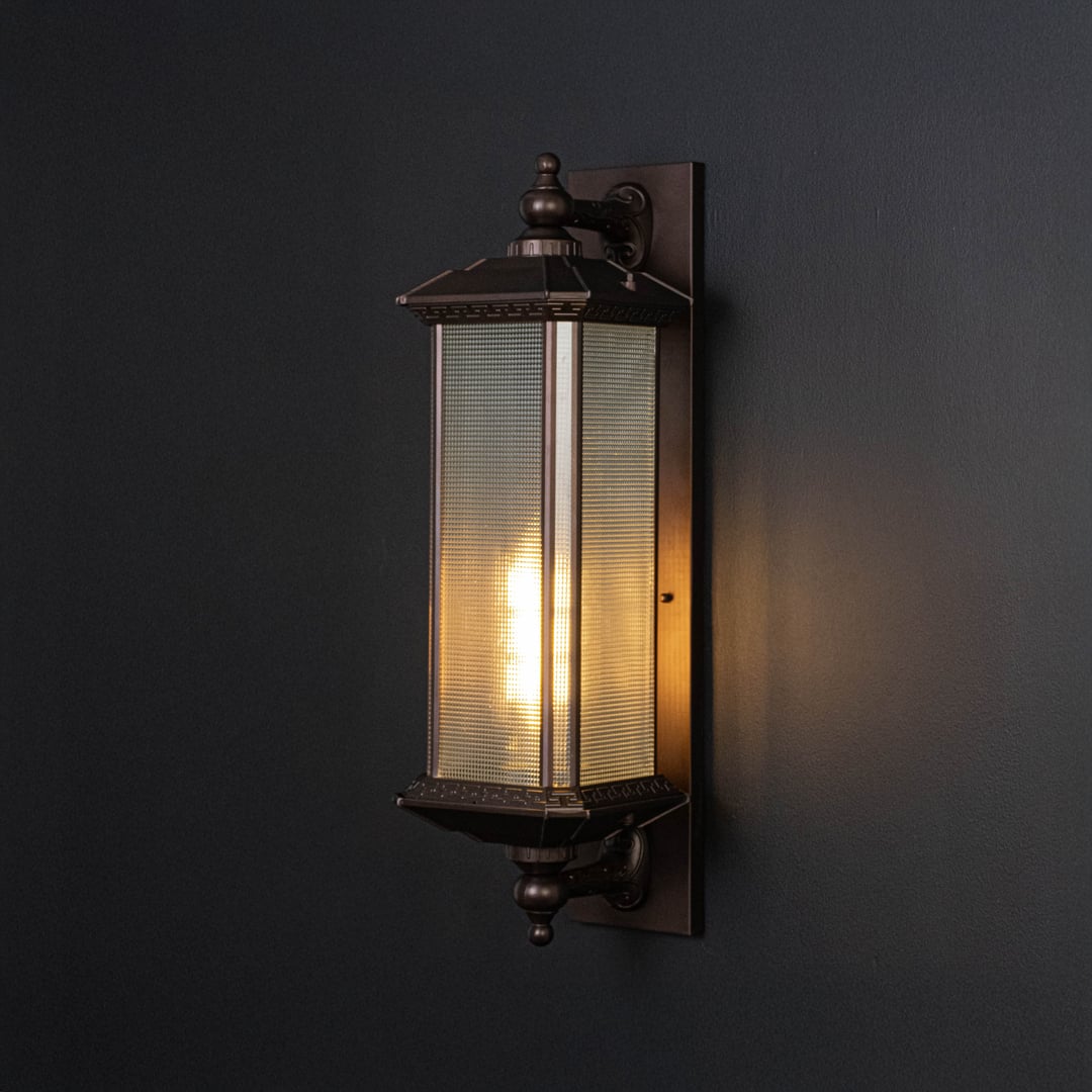 Outdoor bronze wall lantern glowing softly with frosted glass shade creating warm ambience on dark wall.