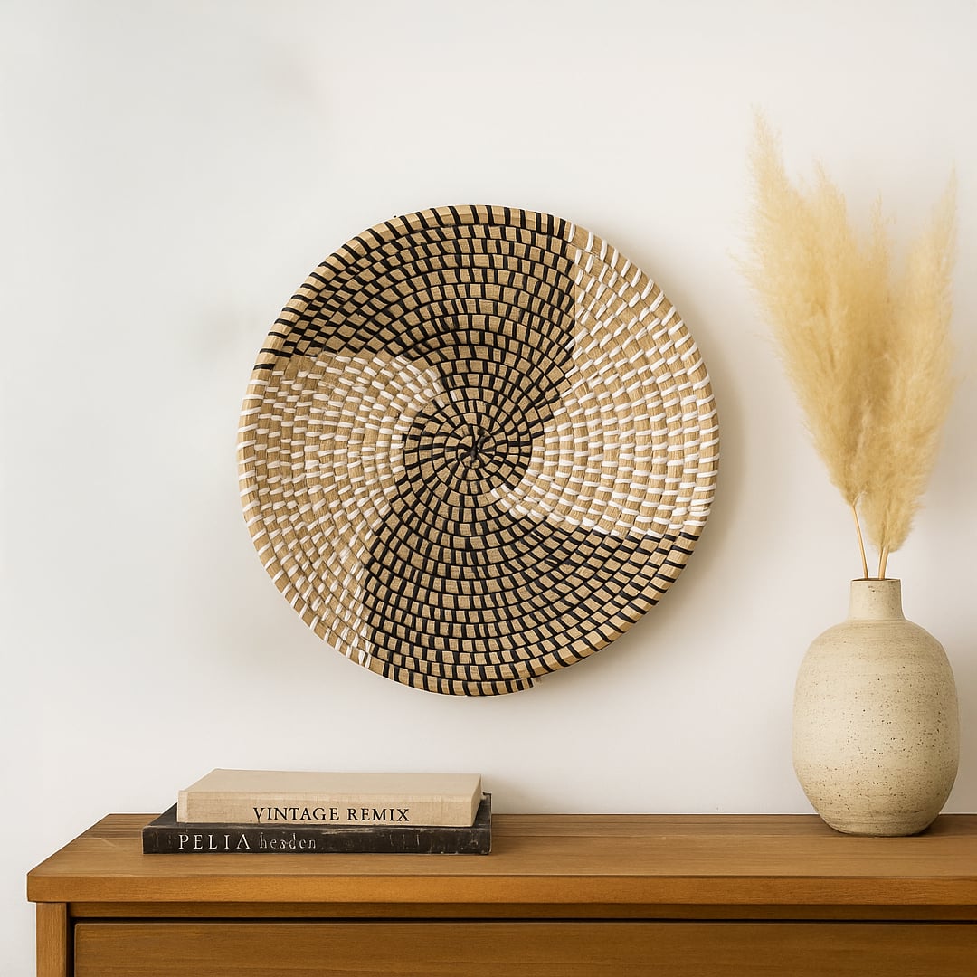 A round woven wall basket with a black and white spiral pattern hangs above a wood cabinet holding stacked books and a beige vase of pampas grass.