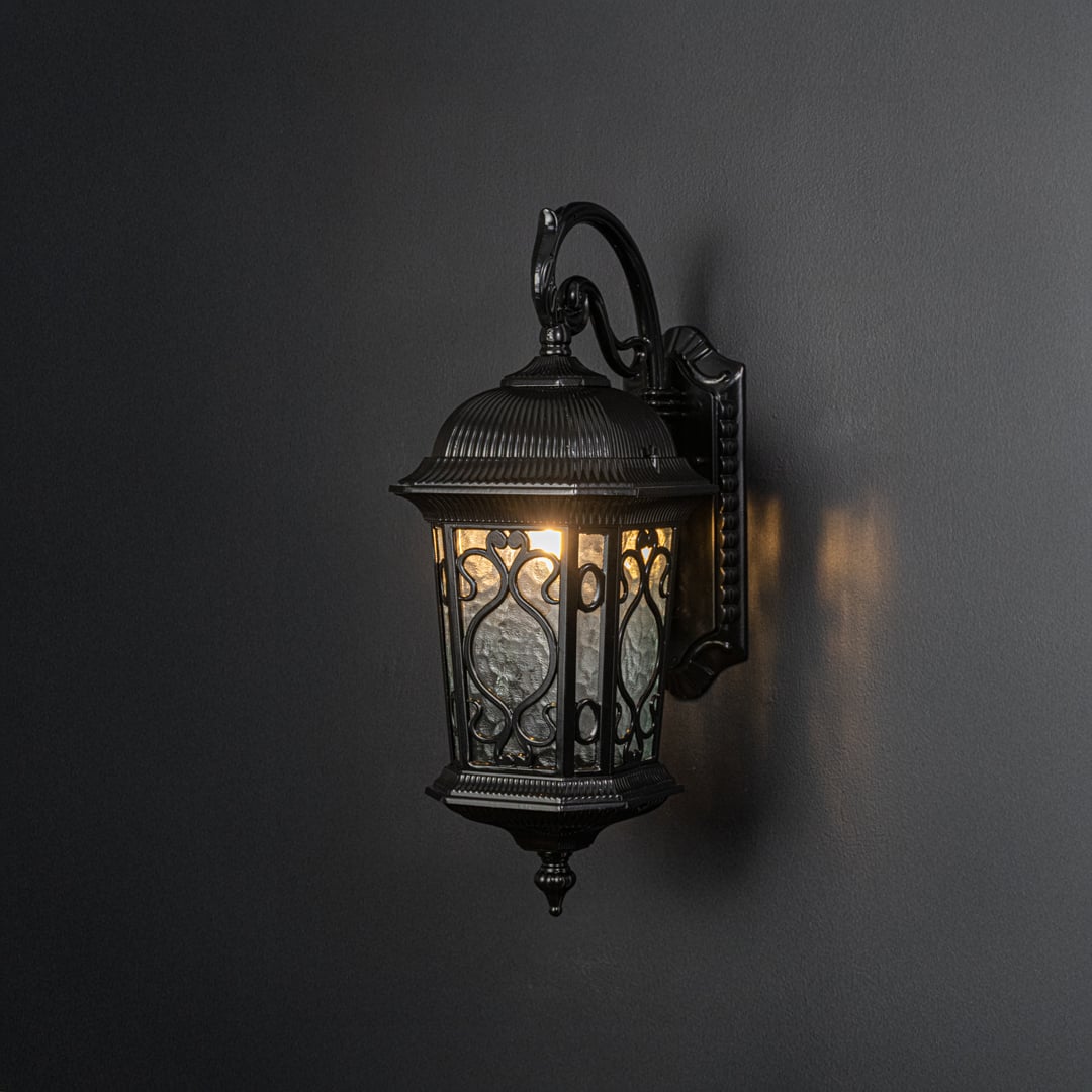 Light on black background features black metal outdoor wall lantern illuminated to emphasize vintage styling and warm light effect.