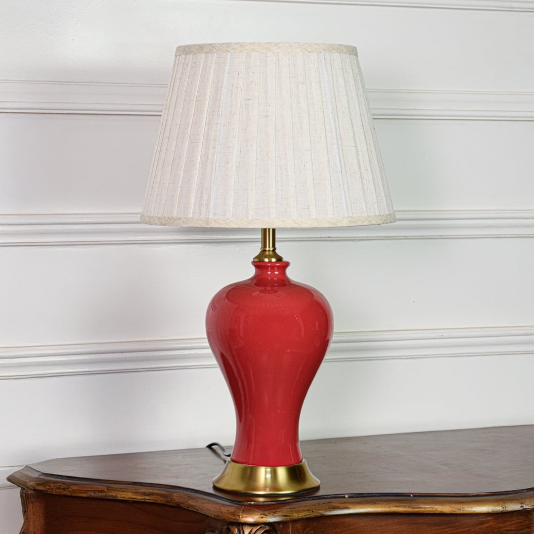 Modern red ceramic table lamp with glossy body gold metal base and pleated fabric shade on soft off white background.
