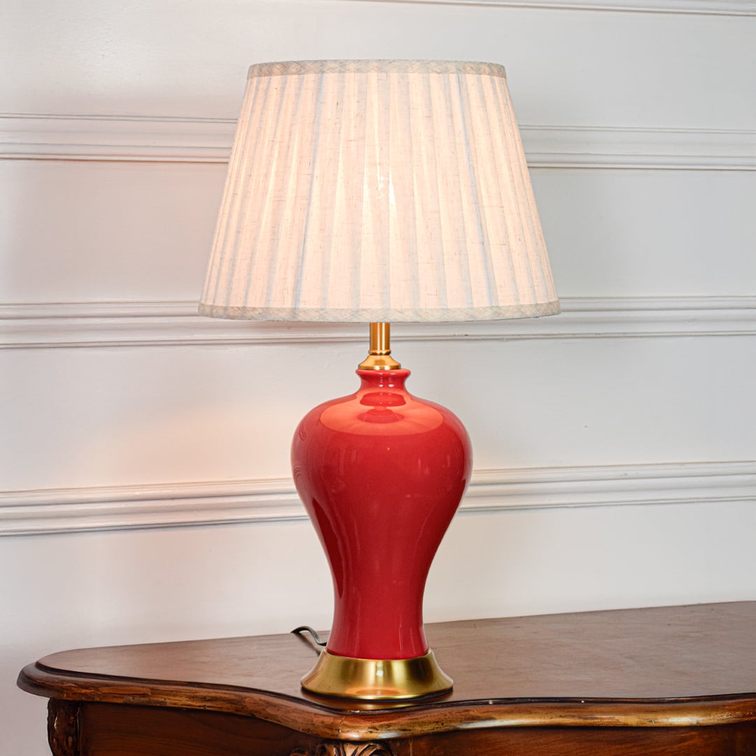 Red ceramic table lamp featuring smooth glossy finish gold metal base and pleated fabric shade displayed on white background.