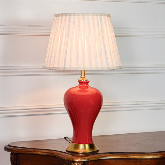 Red ceramic table lamp featuring smooth glossy finish gold metal base and pleated fabric shade displayed on white background.