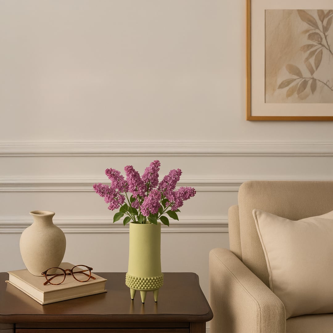 Light green ceramic vase with textured base holding purple flowers placed on wooden side table beside beige sofa.