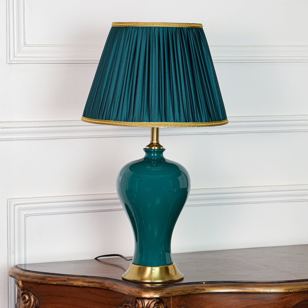 Modern teal ceramic table lamp with glossy body gold metal base and pleated fabric shade placed on soft off white background.