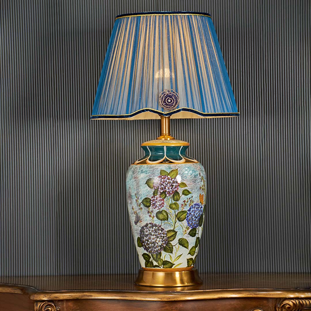 Traditional blue ceramic table lamp turned on with ornate floral artwork and pleated blue shade standing out against a dark background for classic decor style.
