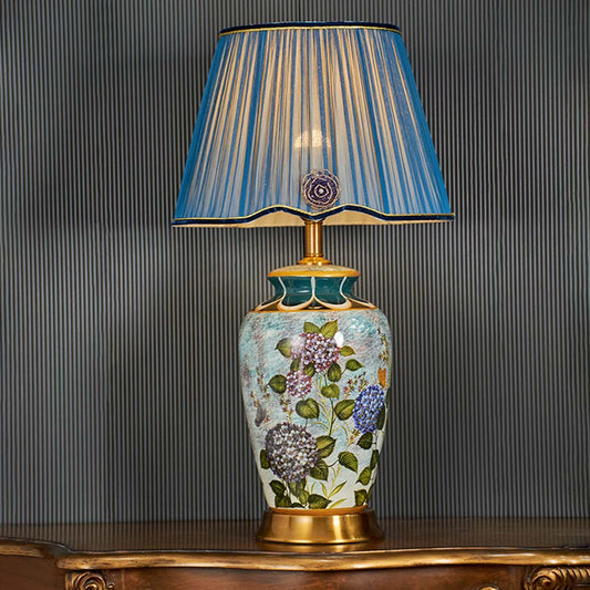 Traditional blue ceramic table lamp turned on with ornate floral artwork and pleated blue shade standing out against a dark background for classic decor style.